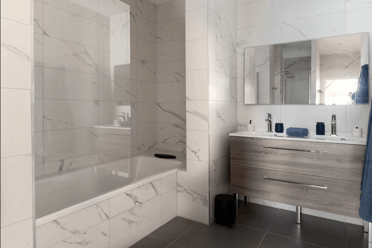 A modern bathroom interior featuring white marble-look wall tiles, a built-in bathtub with a glass shower screen, a floating wood vanity with a white sink, and a large mirror.