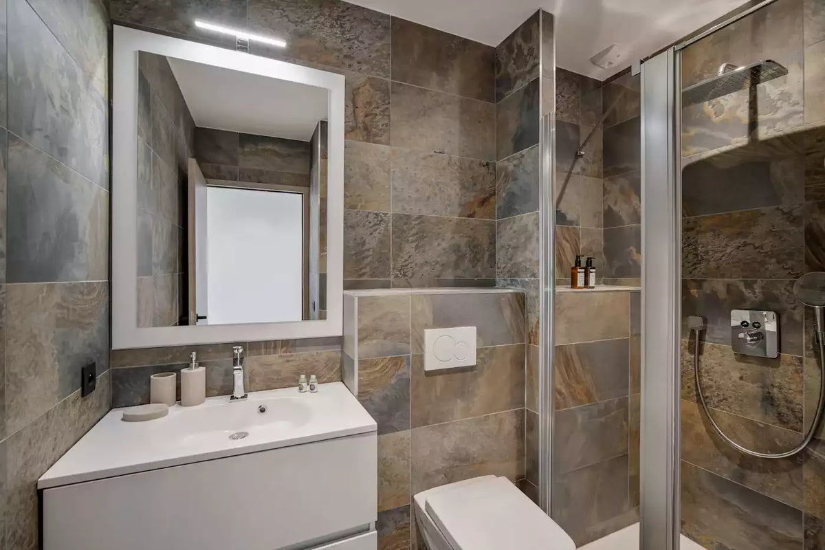 Modern bathroom interior with stone-look tiled walls, a white vanity and sink, a large rectangular mirror, a toilet, and a glass shower enclosure with chrome fixtures.
