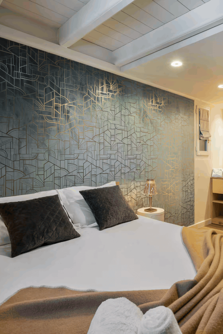 A modern bedroom interior featuring a neatly made bed with white linens, dark accent pillows, and a tan throw blanket. The wall behind the bed has a large geometric metallic pattern in silver and dark gray tones, while the adjacent wall is made of light natural wood paneling.