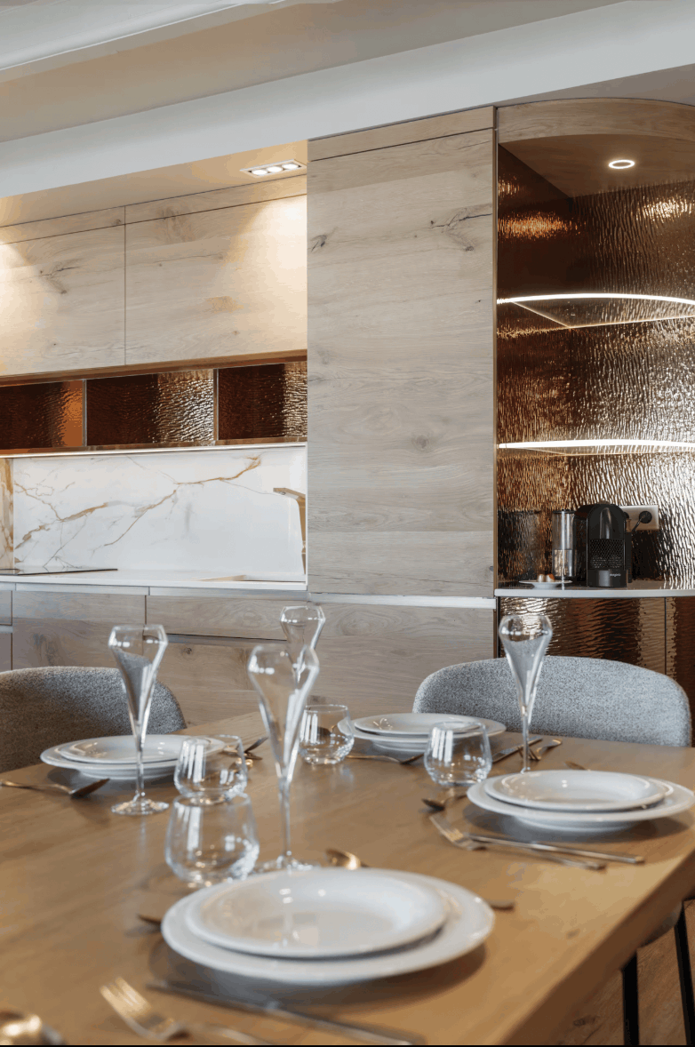 A modern dining table set with white plates, clear glassware, and silver cutlery, set against a contemporary kitchen backdrop with light wood cabinets, a marble backsplash, and textured metallic copper-toned wall panels.