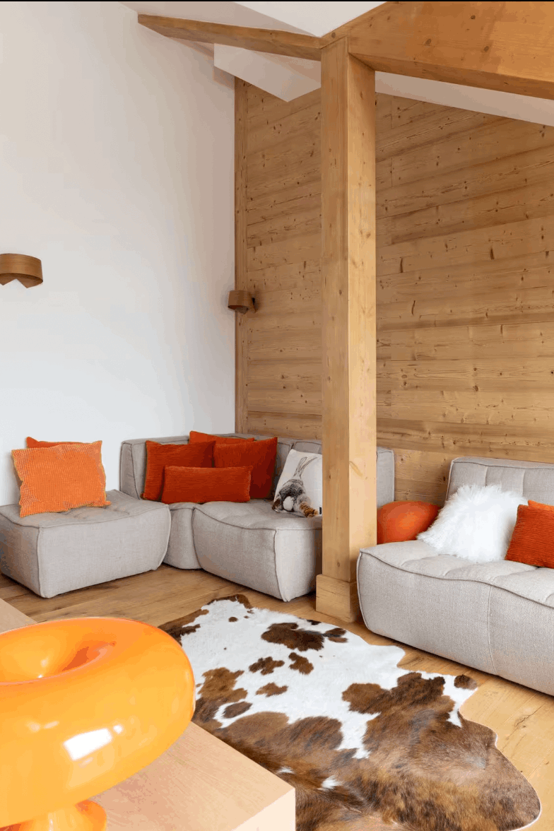 A modern living room with a light gray sectional sofa accented by bright orange and white pillows, set against a wood-paneled wall with exposed wooden beams.
