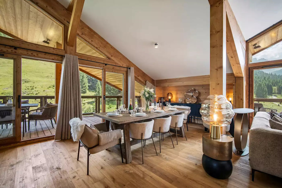 A luxury modern dining room with a long wooden dining table, beige upholstered chairs, a glass globe table lamp, and a cozy sofa, set in a rustic wooden chalet with a mountain view.