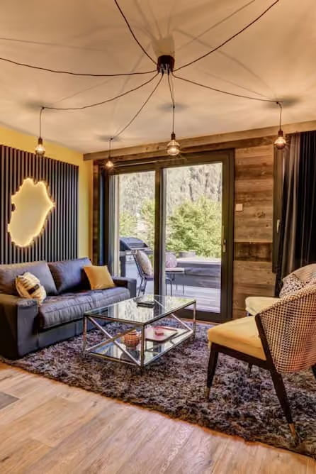 A modern living room interior with a dark gray sofa, glass coffee table, textured rug, wood flooring, yellow accent wall, patterned orange wall, and large windows with outdoor greenery visible.