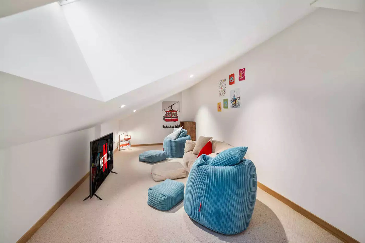 A modern home theater setup in a white-walled attic room, featuring a large blue corduroy bean bag chair, matching ottomans, a beige sofa with red and blue cushions, a TV on a stand, and a small shelving unit.