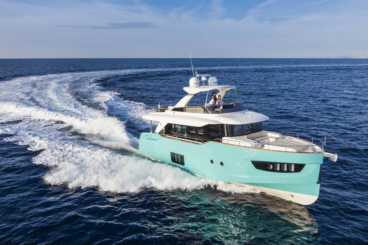 A luxury motor yacht in a light turquoise and white color combination, captured from an aerial perspective as it cuts through deep blue ocean water, leaving a white wake behind it.