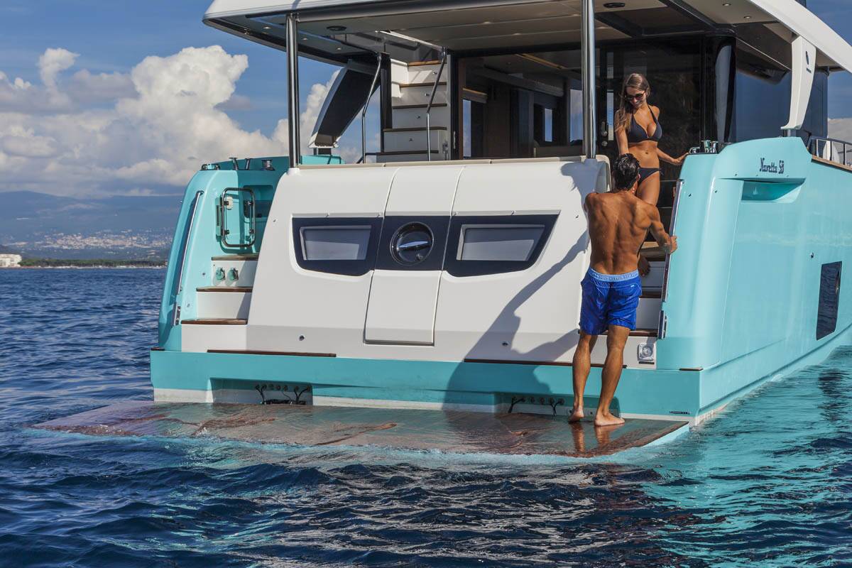 A turquoise and white luxury motor yacht on open blue water, with two people standing on the rear deck and a sliding platform extending into the sea.