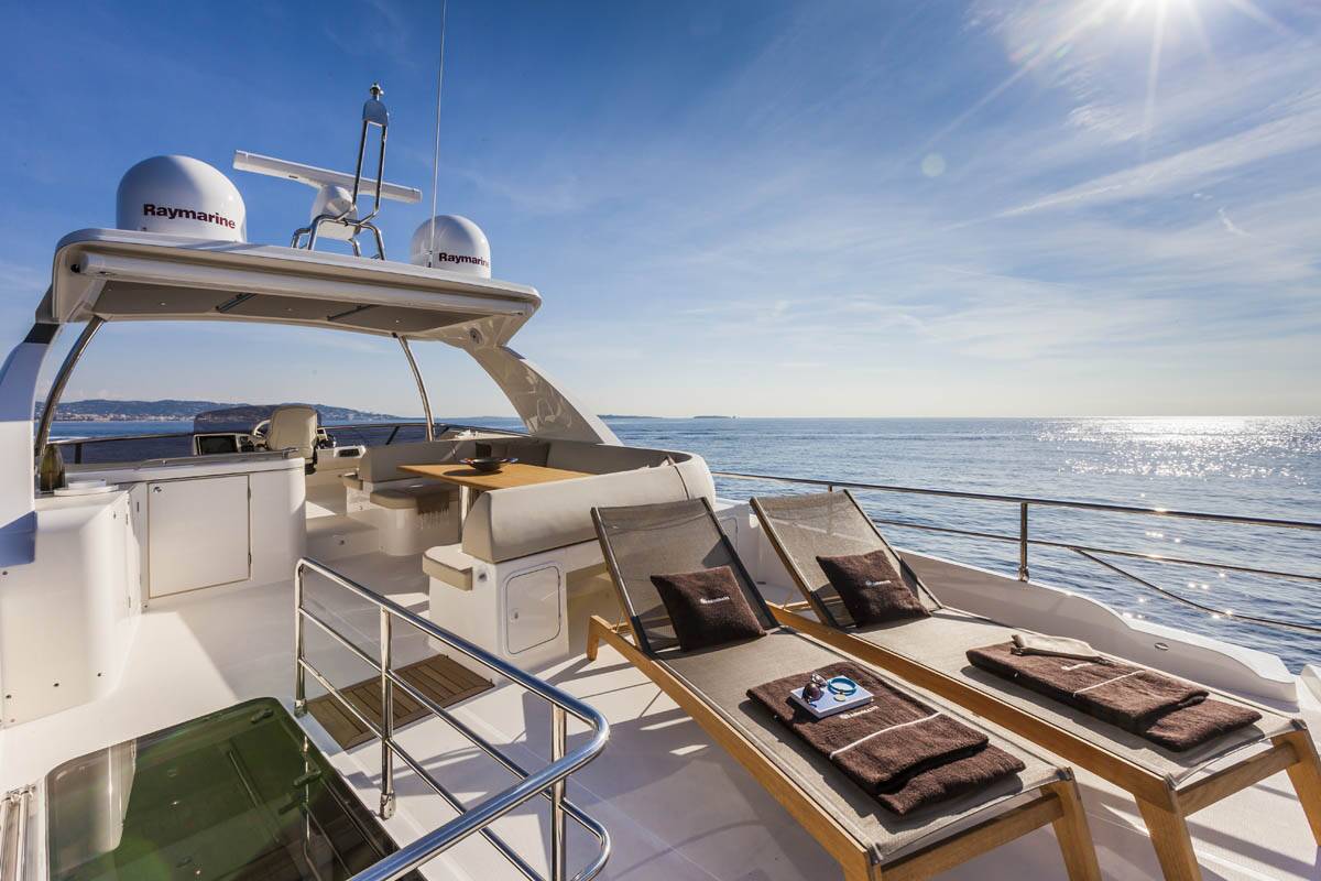 A luxury yacht deck featuring two wooden sun loungers with dark brown towels, a radar arch with Raymarine domes, and an open view of the blue ocean under a bright sun.