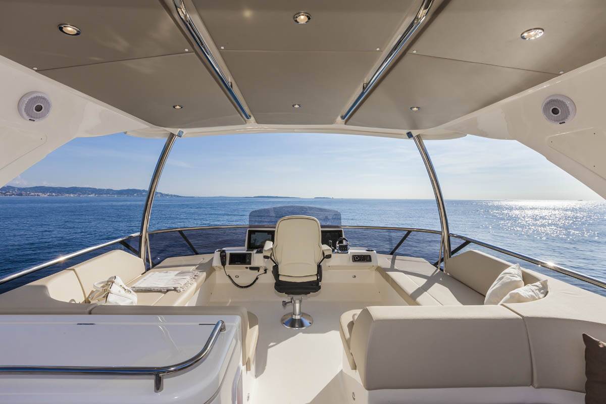 The helm station and cockpit of a luxury yacht, featuring cream-colored upholstery, a central captain's chair, navigation consoles, and a panoramic view of the blue ocean under a clear sky.