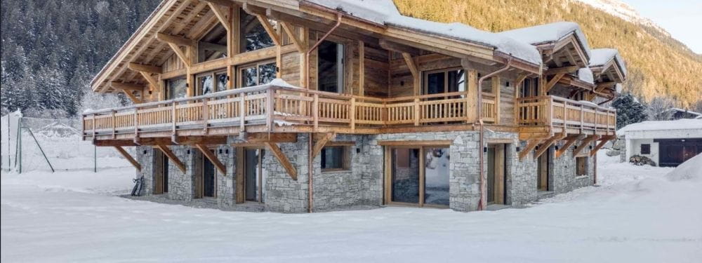 A large wooden chalet-style mountain house with snow-covered roofs and balconies, set in a snowy alpine landscape with trees and hills in the background.