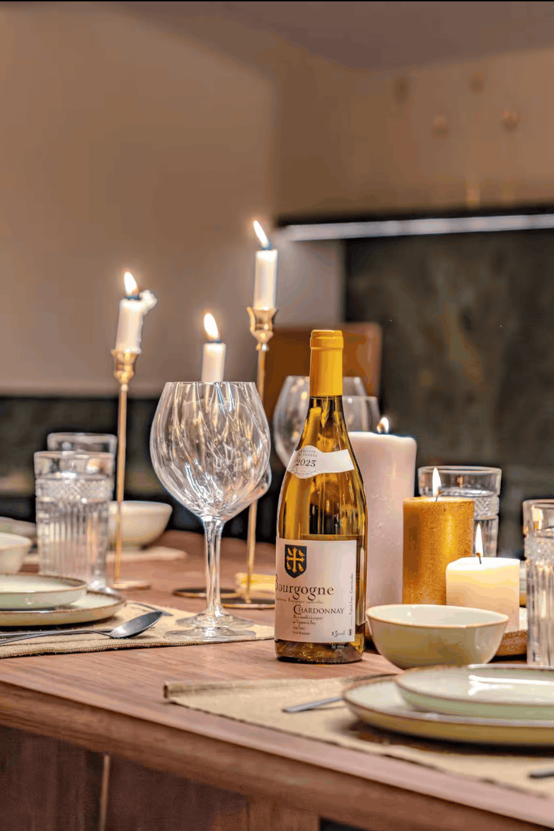 A styled dining table set for guests with wine glasses, plates, candles, and a bottle of wine in a warm, rustic kitchen setting.