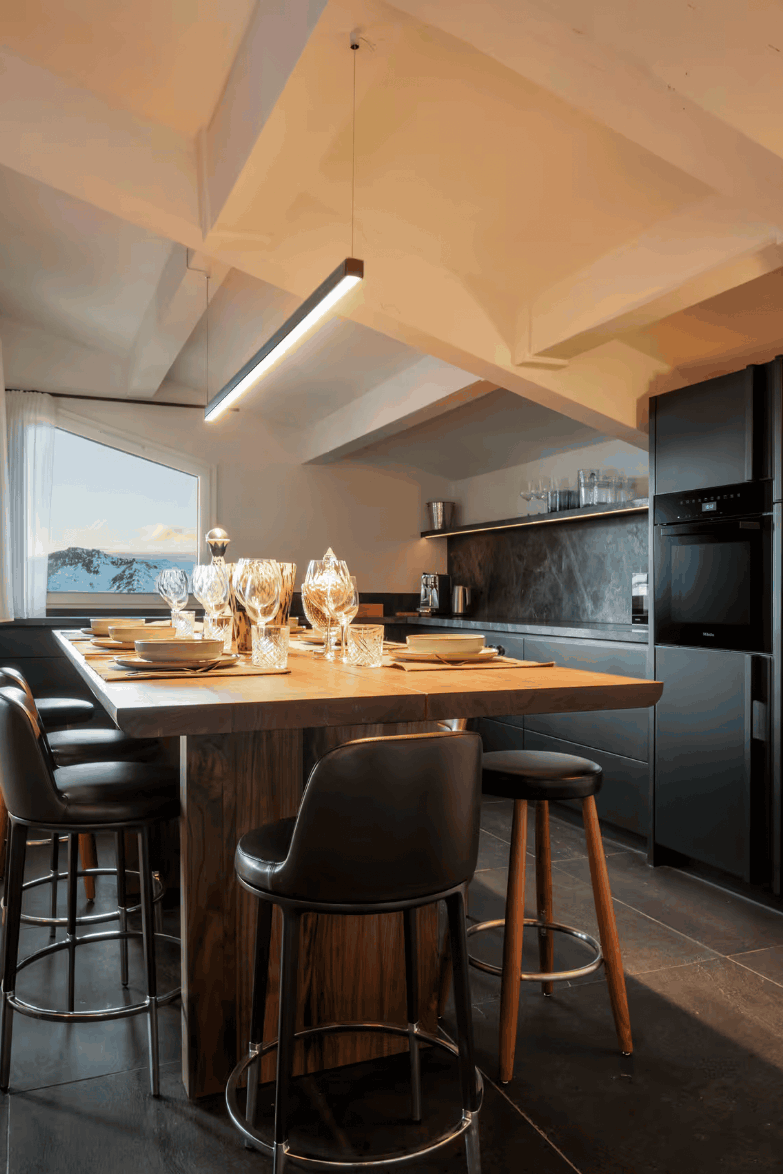 A modern kitchen island with a wooden countertop and black bar stools, set in a spacious kitchen with dark appliances, a high slanted ceiling, and a window showing a mountain view.