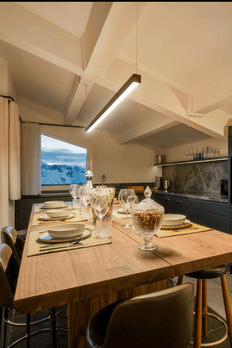 A modern kitchen and dining area with a tall wooden island table set with place settings and bar stools, dark cabinetry, a slanted ceiling with pendant lights, and a window showing an outdoor view.