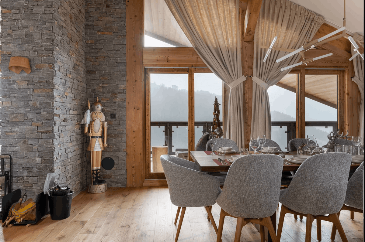 A modern dining room with a wooden table and gray upholstered chairs, set in a rustic chalet-style interior with stone walls, wood beams, and large windows overlooking a mountain view.