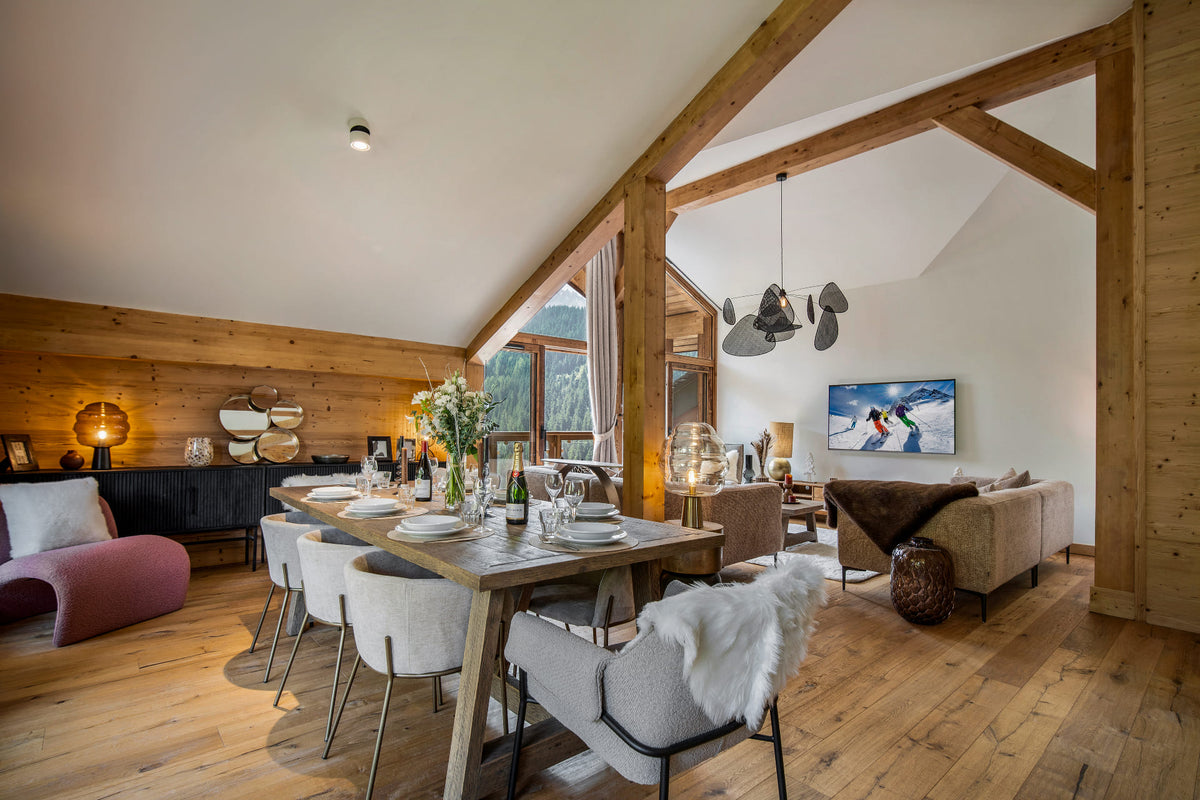 A modern ski chalet dining and living room featuring a rustic wooden dining table with white chairs, a large TV on the wall, and a cozy sofa in the background, all set under a sloped ceiling with exposed wooden beams.