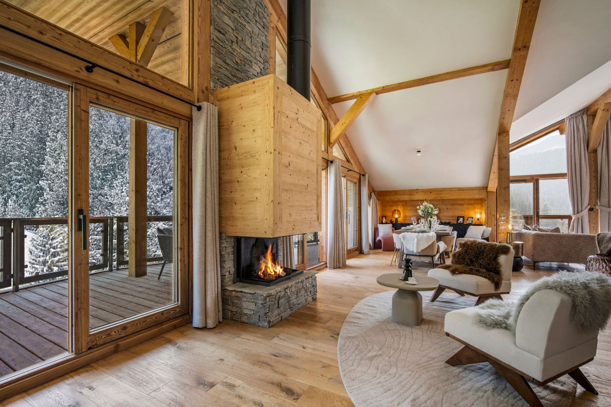 A luxurious ski chalet living room featuring a large wood-burning fireplace, light oak wood floors, high vaulted ceilings with exposed wooden beams, and a cozy seating area with modern armchairs and a round rug.