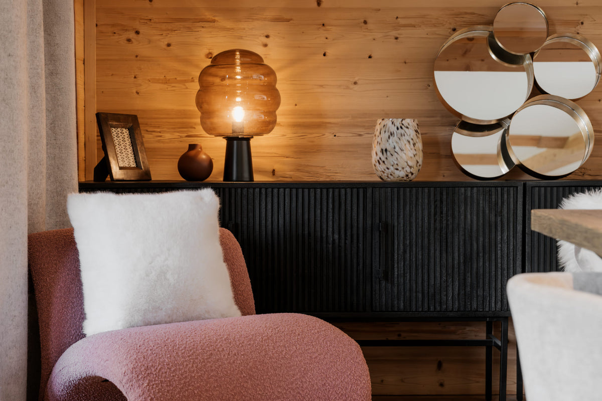 A cozy interior scene with a pink upholstered armchair holding a fluffy white throw pillow, set against a wood-paneled wall with a lit amber table lamp, decorative vase, mirrors, and a dark ribbed cabinet.
