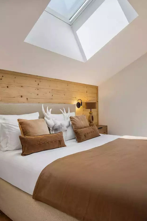 A modern bedroom interior featuring a neatly made bed with white linens and brown pillows, set against a light wood headboard wall. A skylight is visible above the bed, and a wall-mounted TV is shown on the right.