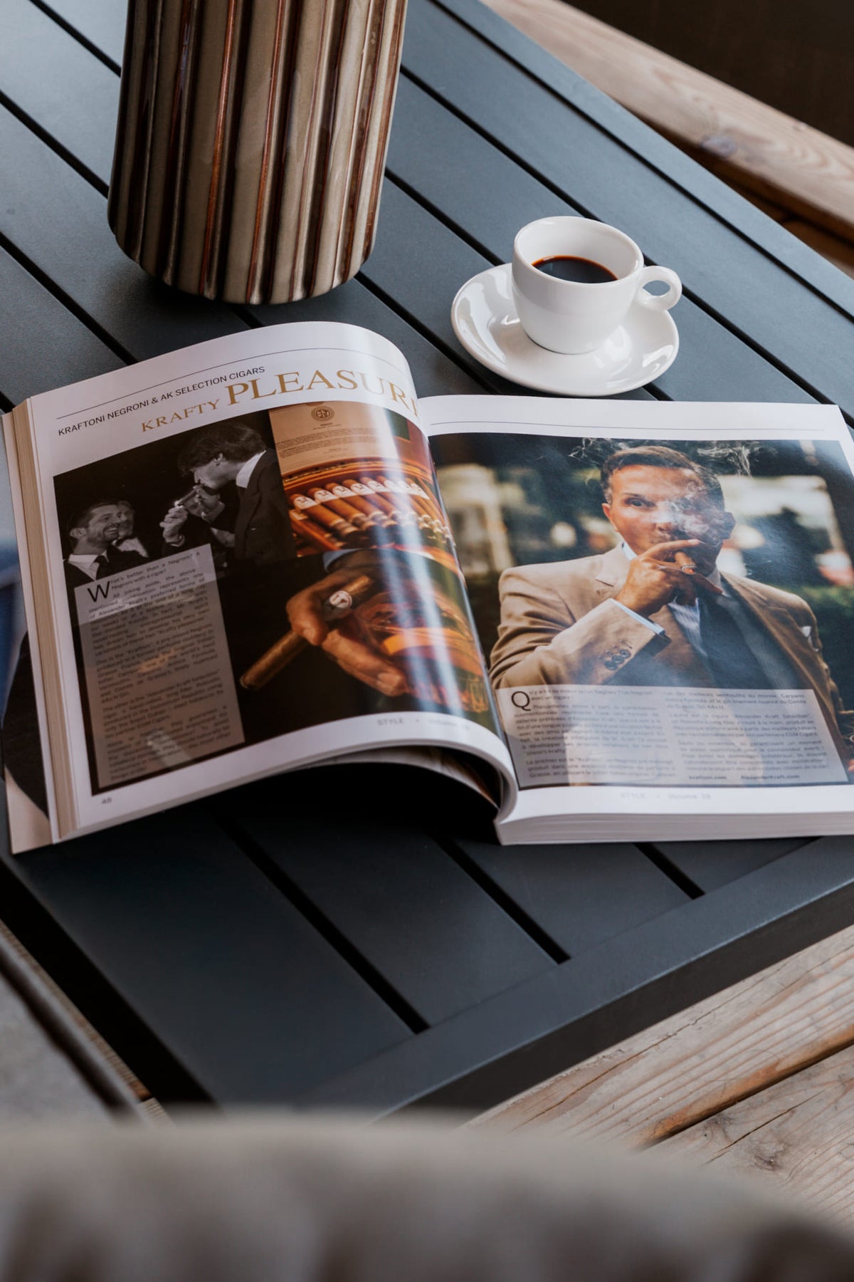 An open magazine spread lies on a dark slatted table next to a cup of coffee on a saucer, with a striped cylindrical container in the background.