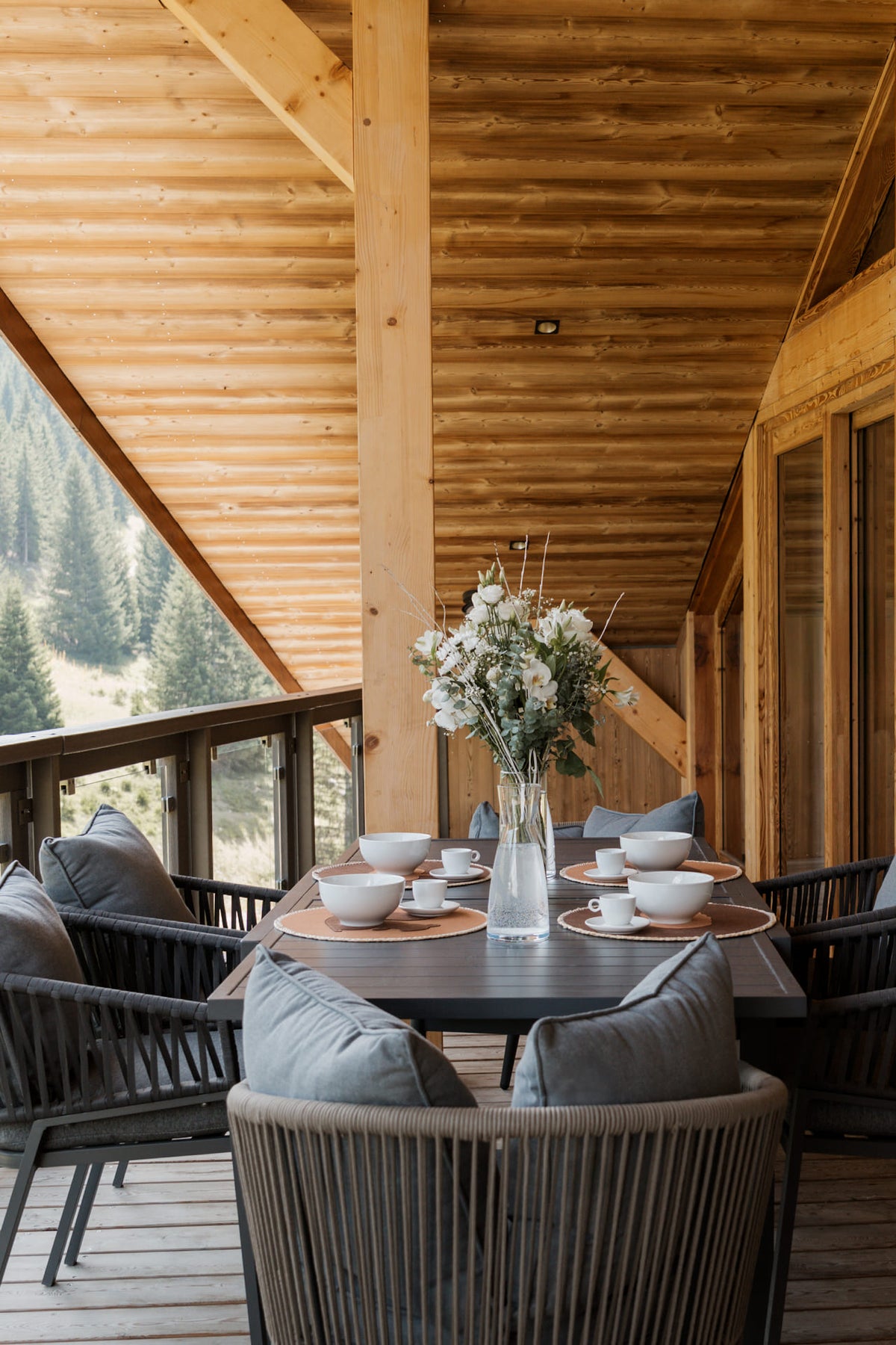 A dining set on a wooden deck with a black rectangular table, gray woven chairs with cushions, white ceramic bowls, and a clear glass vase holding white flowers. The setting is on a wooden chalet balcony overlooking a forest.