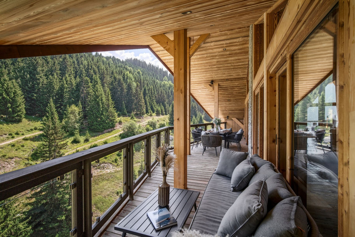 A modern wooden deck with a built-in outdoor sofa, coffee table, and large glass windows overlooking a lush green forest valley.