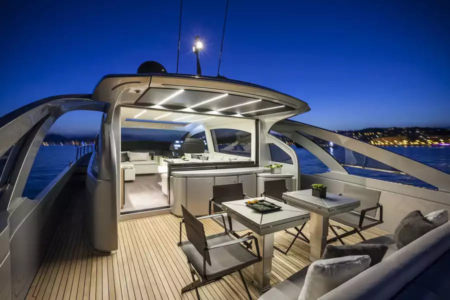 Luxury yacht outdoor deck at twilight with a dining area, bar seating, and open upper deck cabin, surrounded by water and distant city lights.