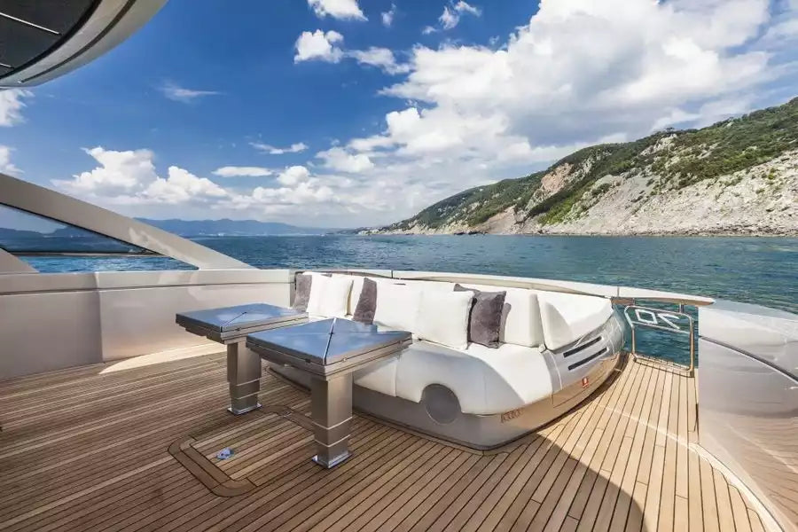 A luxury yacht outdoor deck featuring a white curved lounge sofa with gray pillows, a matching square bar table, and a wooden teak deck, overlooking a scenic blue sea and green rocky coastline under a partly cloudy sky.