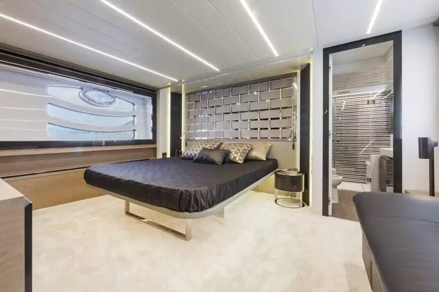 A luxury modern bedroom featuring a low-profile platform bed with a dark gray bedding set, a metallic grid headboard, and a large window looking out onto a yacht deck.