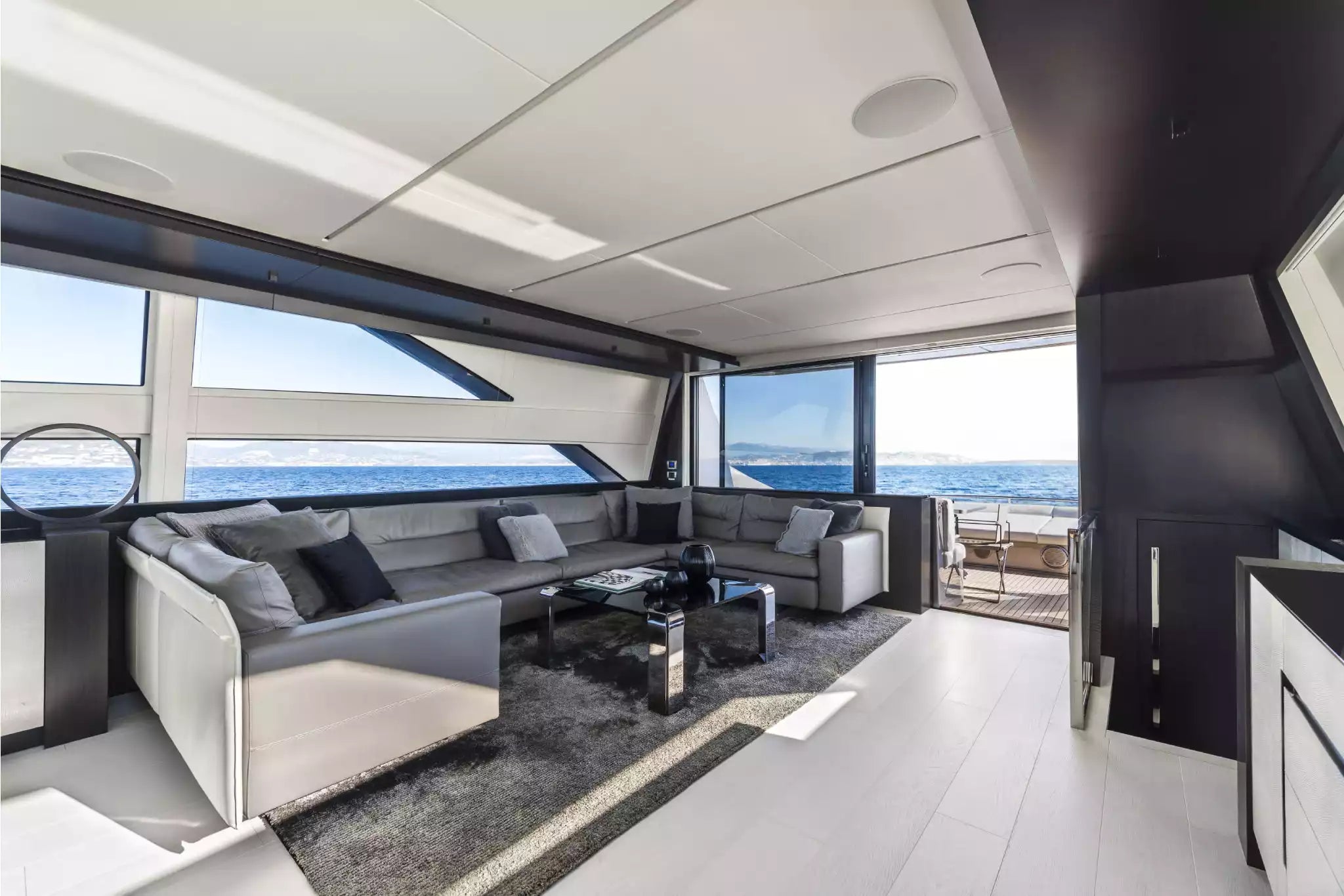 A luxury yacht or boat interior featuring a modern U-shaped gray sofa with black accent pillows, a dark wood coffee table, a gray rug, and large windows offering a view of the blue ocean.