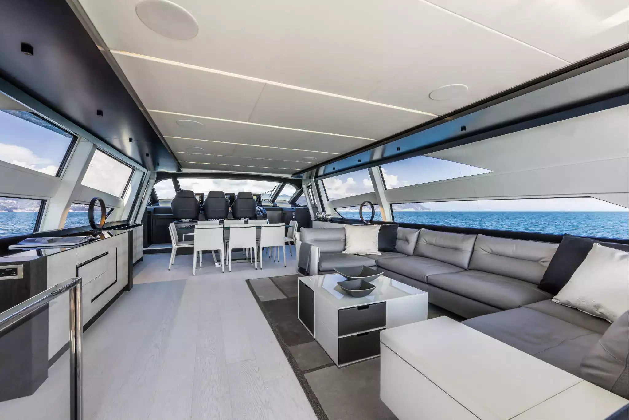 Modern yacht interior with a spacious living area featuring a gray sectional sofa, white ottoman, glass coffee table, dining set, and large windows overlooking the blue ocean.