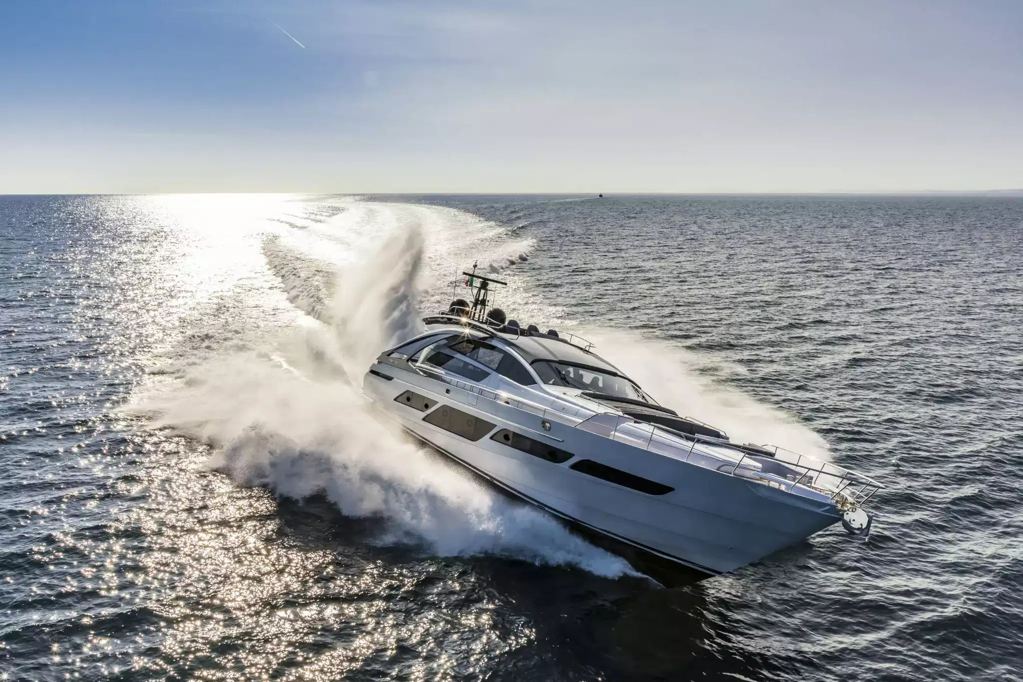 A luxury motor yacht speeds across open ocean water, creating a large white wake. The yacht is white with dark tinted windows and metallic trim, photographed from an aerial perspective under a clear blue sky.