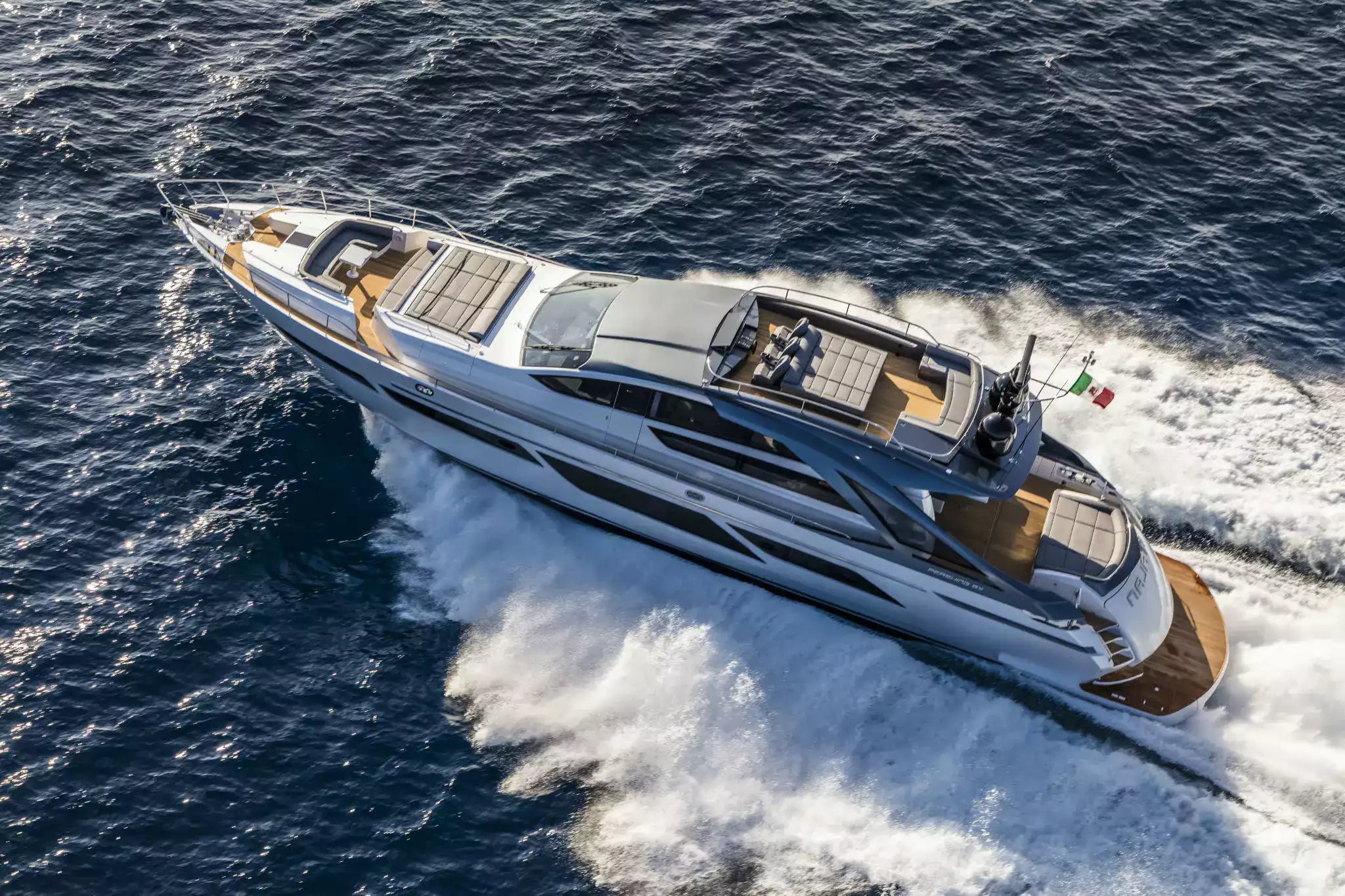 An aerial view of a luxury motor yacht cutting through deep blue ocean water, creating a white wake. The yacht features a white and gray exterior with warm brown wood accents on the deck and bow.