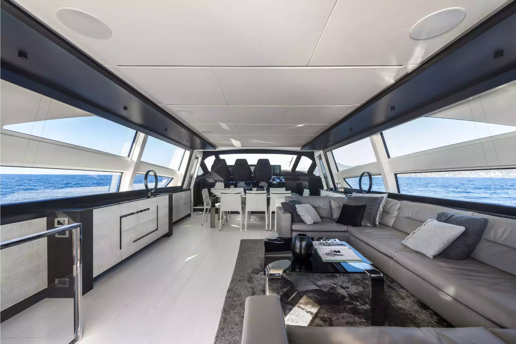 A luxury yacht interior featuring a spacious salon with a gray sectional sofa, black coffee table, and a galley kitchen area with large windows overlooking the blue ocean.