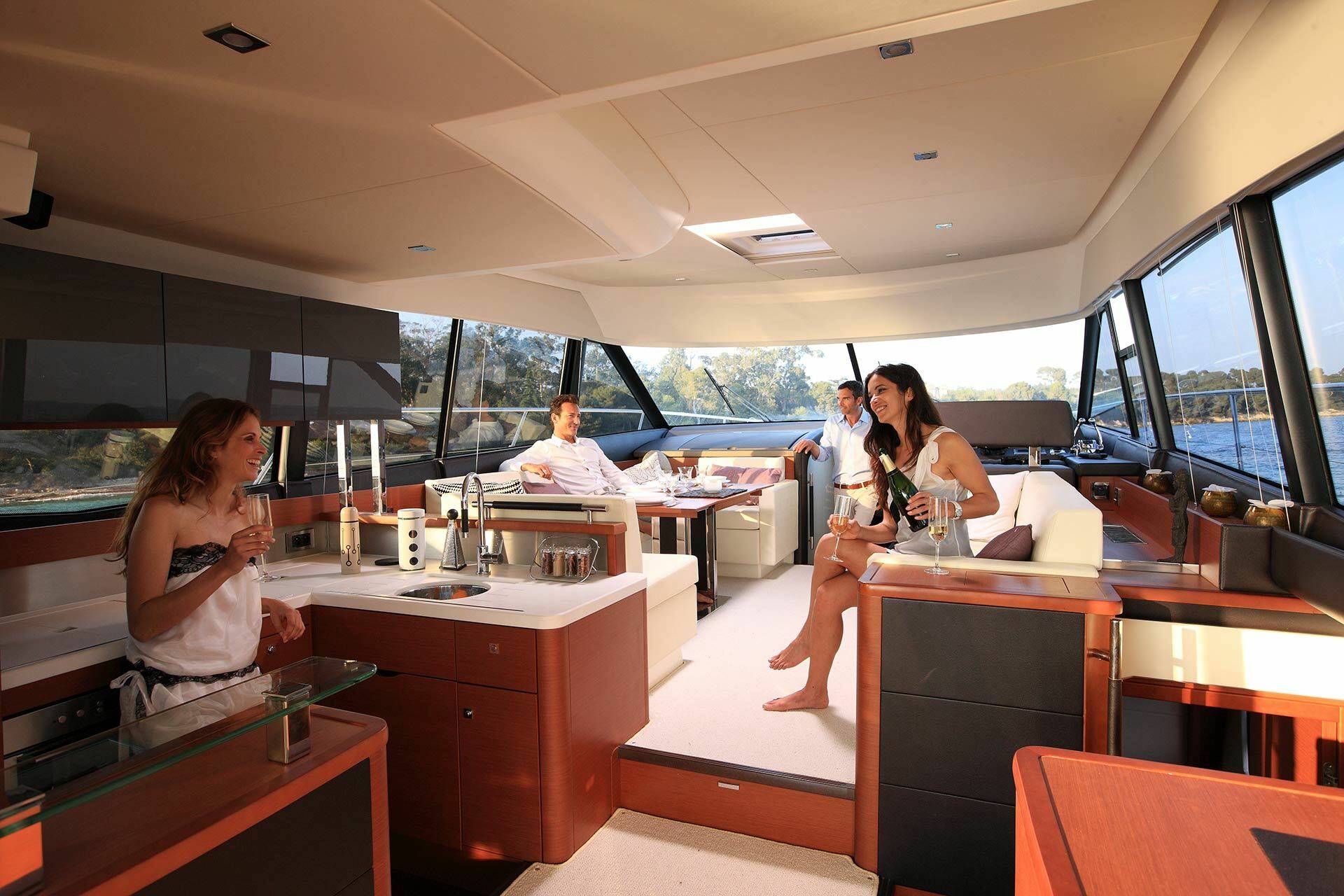 A group of people relaxing inside a luxury yacht cabin with wood-look furniture, white seating, large windows, and views of water and trees outside.