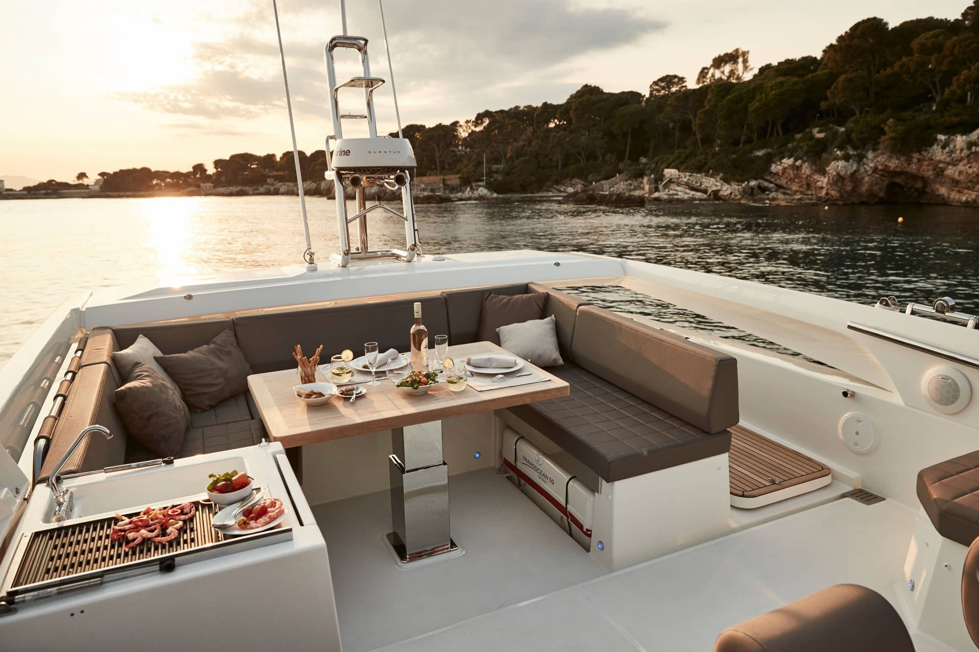 Yacht interior with dining table set for a meal, surrounded by comfortable seating and a scenic view of water and trees.