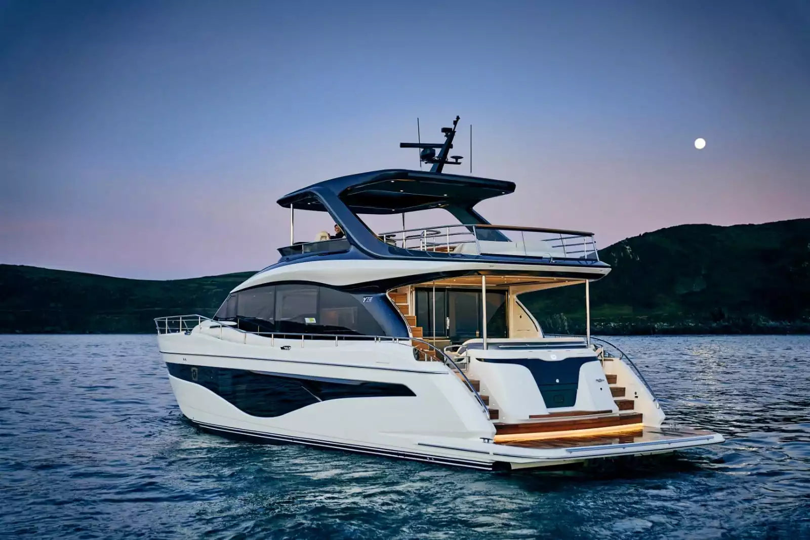 A luxury motor yacht with a white hull and dark blue accents, floating on calm water at dusk with hills and a moon in the background.