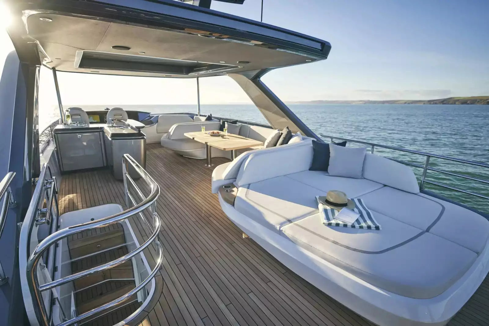 Luxury yacht outdoor deck featuring a large white cushioned lounge daybed, wooden teak flooring, a stainless steel kitchenette, and an open view of the ocean under a clear blue sky.