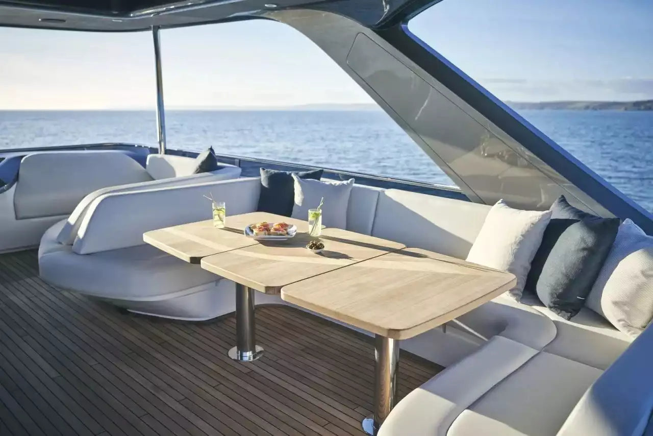 A luxury yacht dining area featuring a light wood table at the center, surrounded by cream and blue upholstered seating with pillows, set against a backdrop of open ocean water under a clear sky.