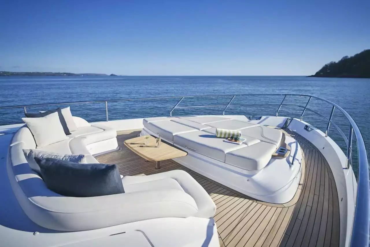 A luxury yacht deck featuring a white curved outdoor lounge sofa with dark blue pillows, a matching white daybed with cushions, a small wooden coffee table, and a wooden teak-look deck, set against a backdrop of blue ocean water and sky.