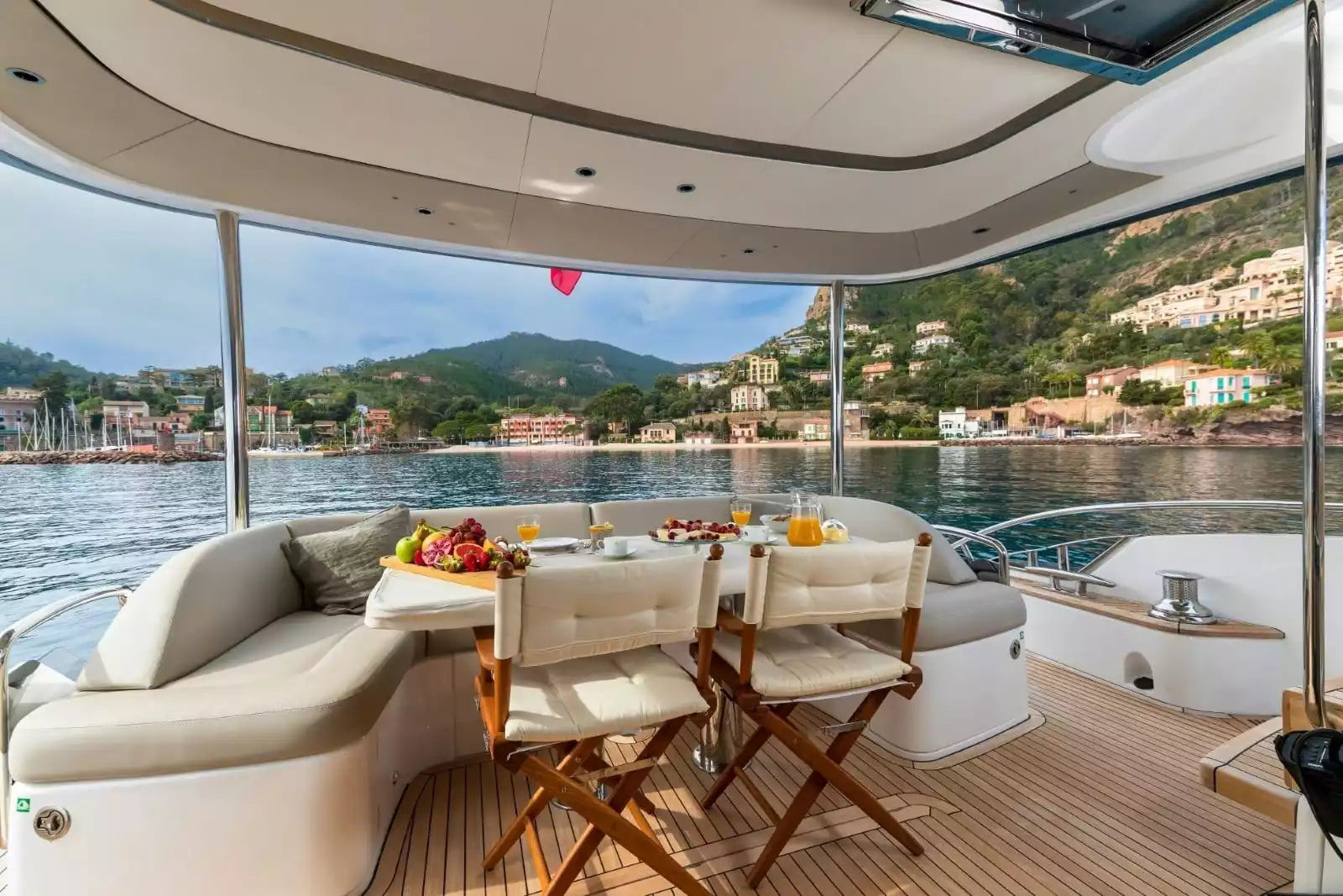 A luxury yacht dining area with a curved cream seating bench, two wooden folding chairs with cream cushions, and a small table set with fruit and drinks, overlooking a scenic waterfront with hills and buildings in the background.