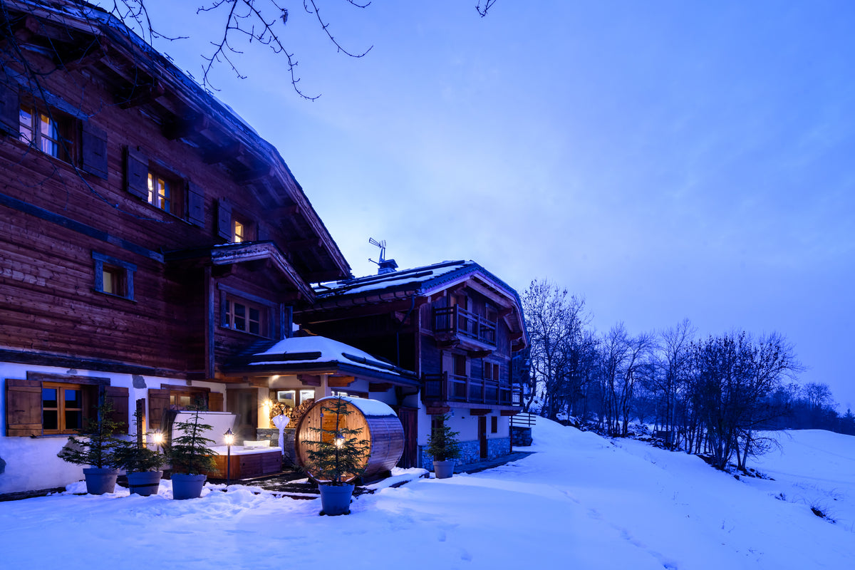 A large traditional wooden chalet in a snowy mountain setting at dusk, with warm lights glowing from the windows and a round wooden barrel structure in the snow in front.