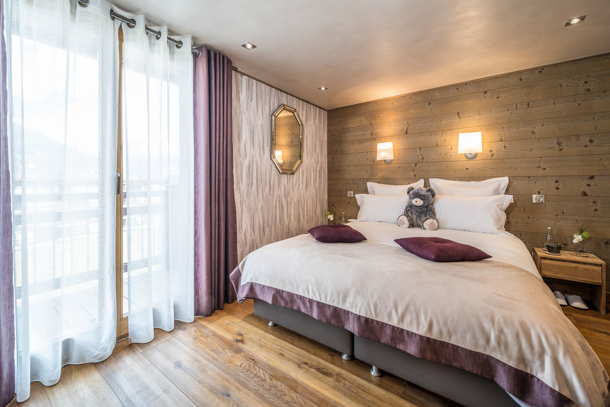 A cozy bedroom interior featuring a neatly made bed with white bedding and dark purple pillows, set against a wood-paneled wall with two wall lamps. Large sheer white curtains and darker purple drapes hang by a glass door on the left, and light wood flooring covers the floor.