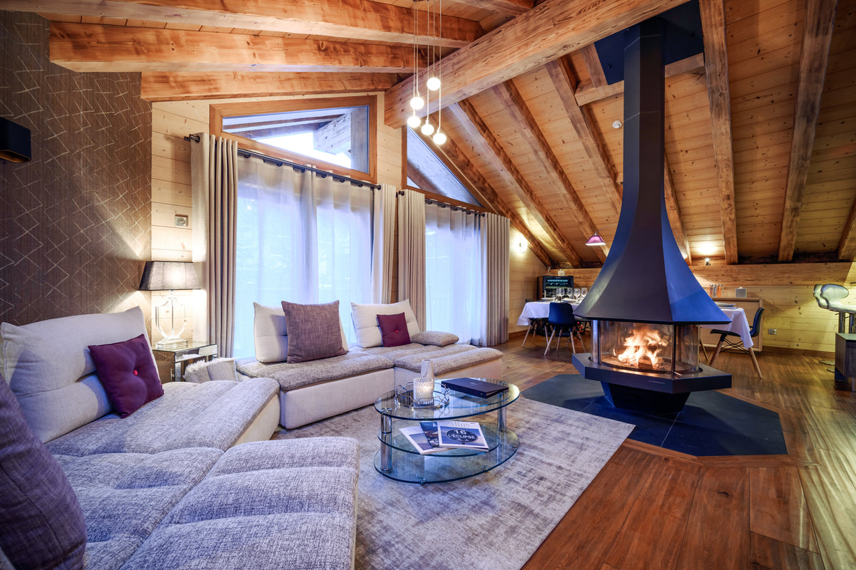 A cozy modern chalet living room featuring a large sectional sofa with purple accent pillows, a glass coffee table, a prominent black wood-burning fireplace with a tall chimney, and a sloped wooden ceiling with exposed beams.
