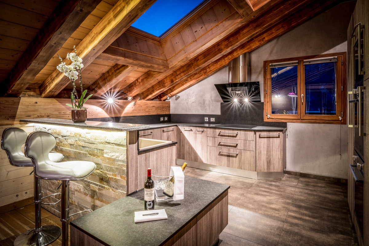 A modern kitchen island in a rustic attic kitchen with wooden ceiling beams, stone backsplash, dark countertops, and two white bar stools.