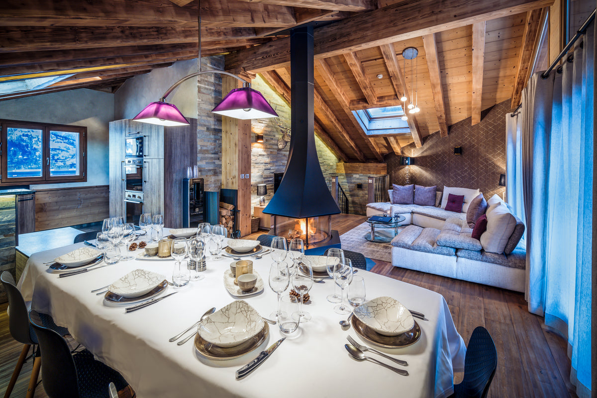 A luxury chalet interior featuring a fully set dining table with white tablecloths and glassware in the foreground, a large black chimney stack, rustic wooden ceiling beams, and a modern sofa in the background.
