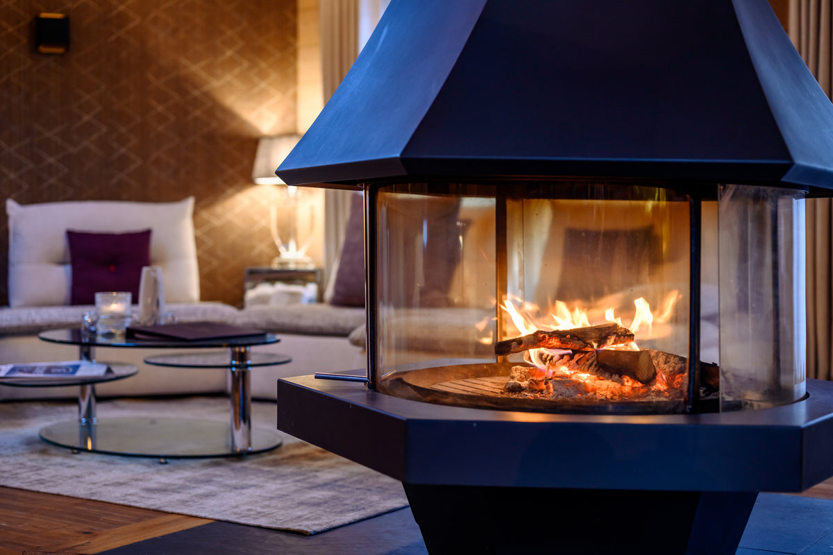 A modern black freestanding fireplace with a glass front and visible flames, set in a warmly lit living room with a sofa and coffee tables in the background.