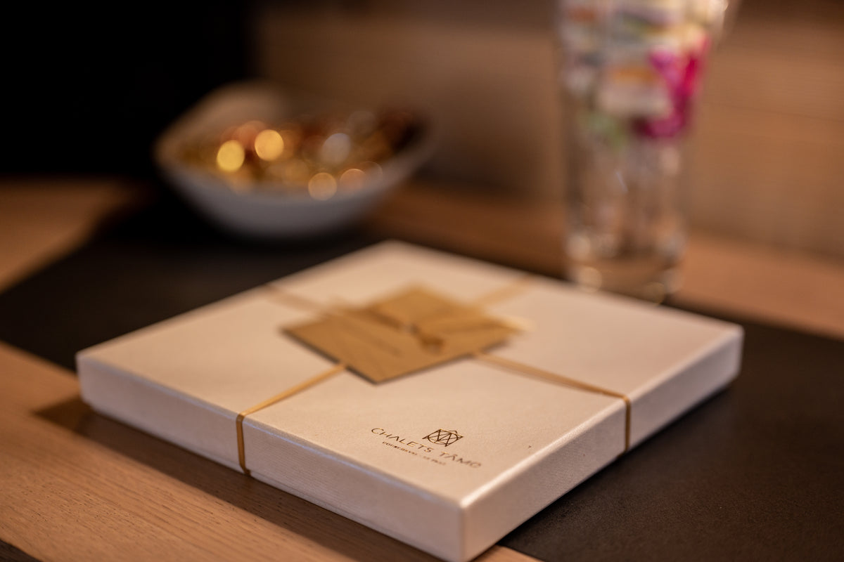 A square cream-colored gift box tied with a thin gold ribbon and a small kraft paper tag, with the brand name 'O'Connell's Piano' printed on the front, set on a dark table runner with a warm, blurred background.