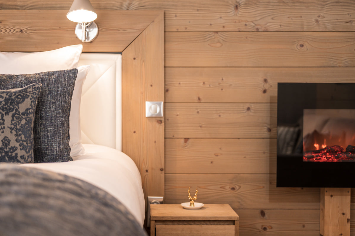 A cozy bedroom corner with a bed dressed in white bedding and dark patterned pillows, set against a light wood-paneled wall. A small wooden bedside table sits between the bed and a wall-mounted electric fireplace showing a glowing fire.