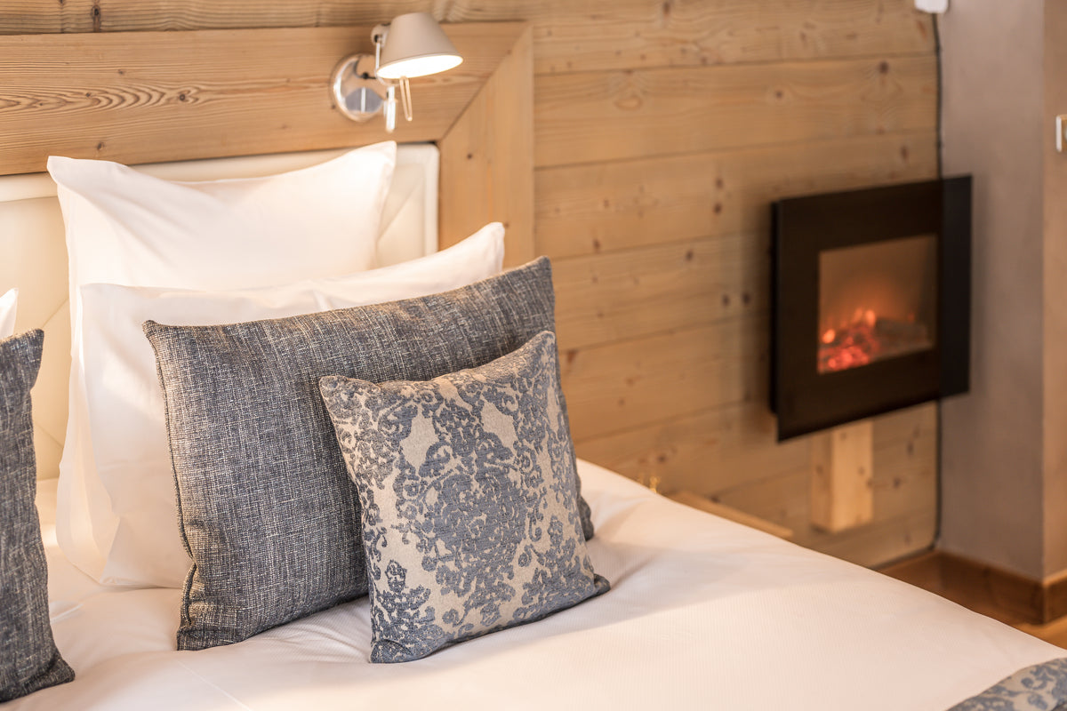 A cozy bedroom scene featuring a neatly made bed with white linens and textured blue-gray pillows, set against a light wood-paneled wall with a wall-mounted electric fireplace.