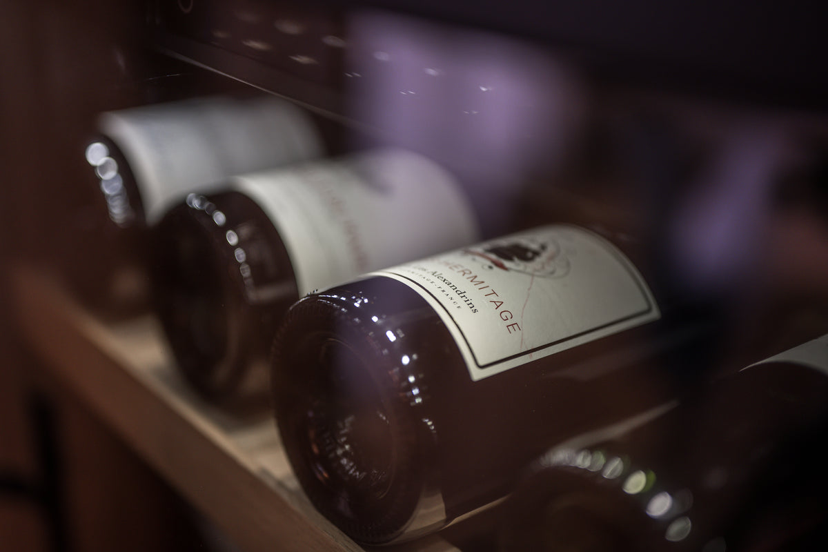 A close-up, shallow depth-of-field shot of a wine bottle on a wooden shelf inside a wine cooler or refrigerator, with other bottles blurred in the background.