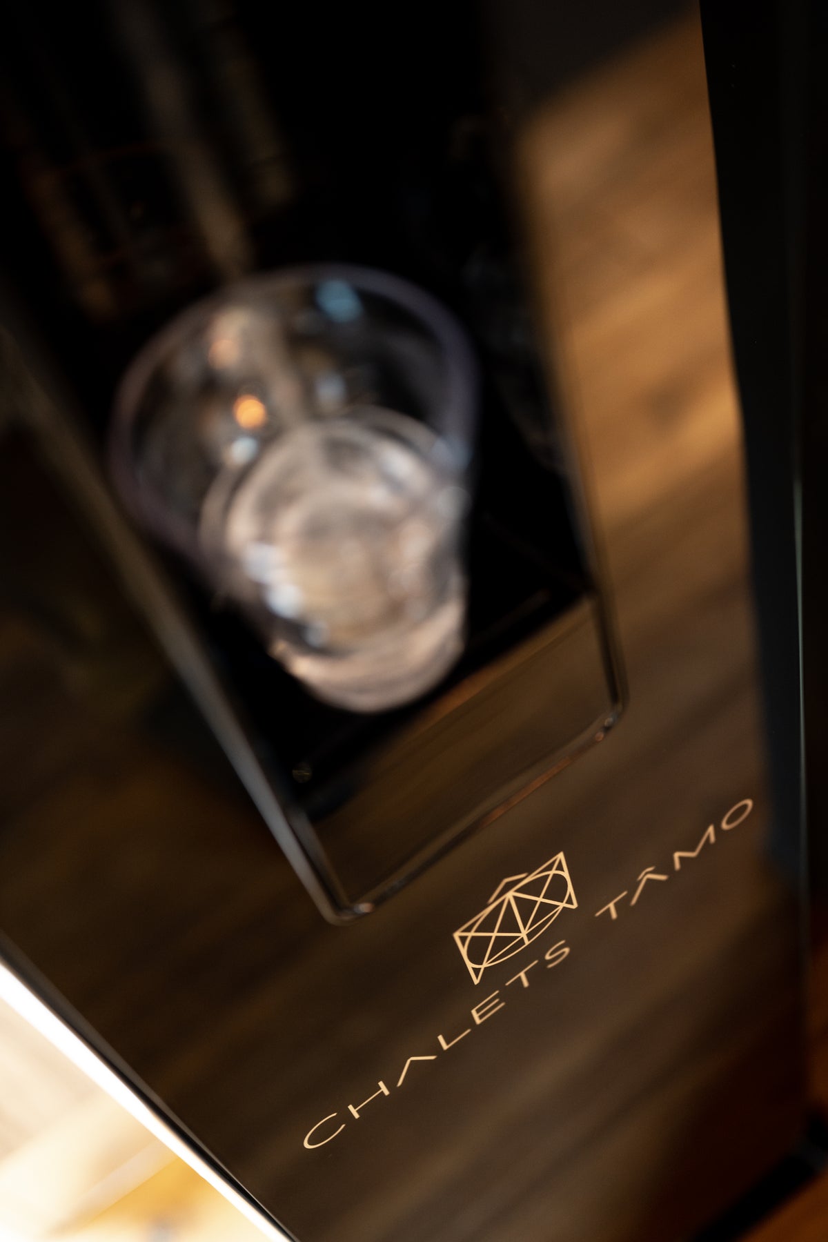 Close-up of a black Chalets Tamo water dispenser with a clear glass cup under the water outlet, on a dark wooden surface.